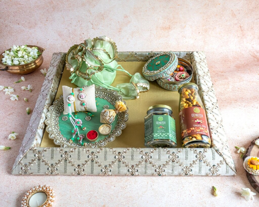Floral Art presents A decorative tray with jewelry, bangles, two jars of snacks, a green pouch, a small ornate box, and scattered flowers on a light surface.