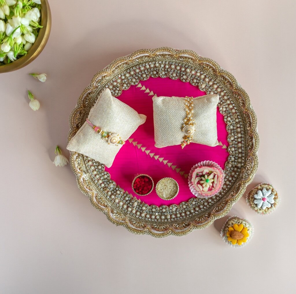 Floral Art presents A decorative tray with two cushion displays holding rakhis, surrounded by small bowls of flowers and colored powder on a pink and gold background.