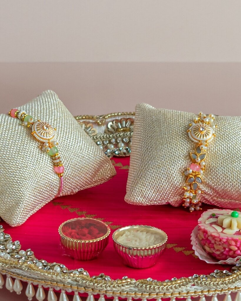 Floral Art presents A decorative tray with two rakhis on cushions, small bowls of red powder and rice, and an Indian sweet, set on a pink fabric background.
