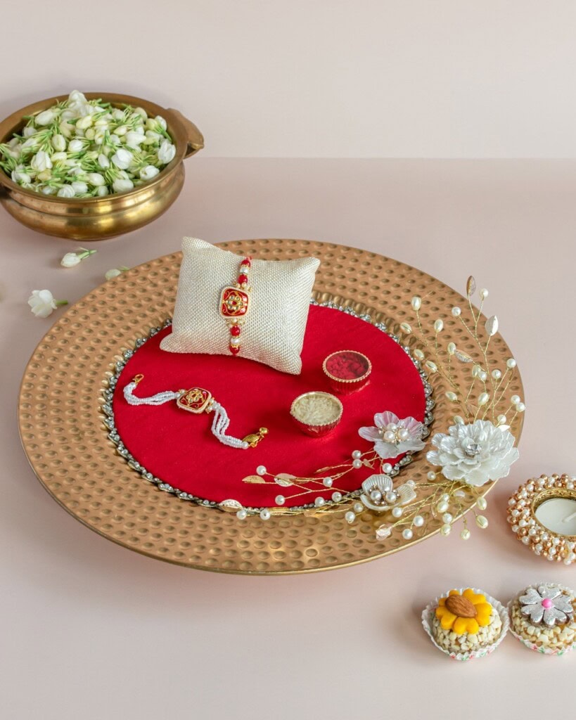 Floral Art presents A decorative plate with a rakhi, small gift box, rings, artificial flowers, sweets, and a bowl of jasmine flowers on a light surface.