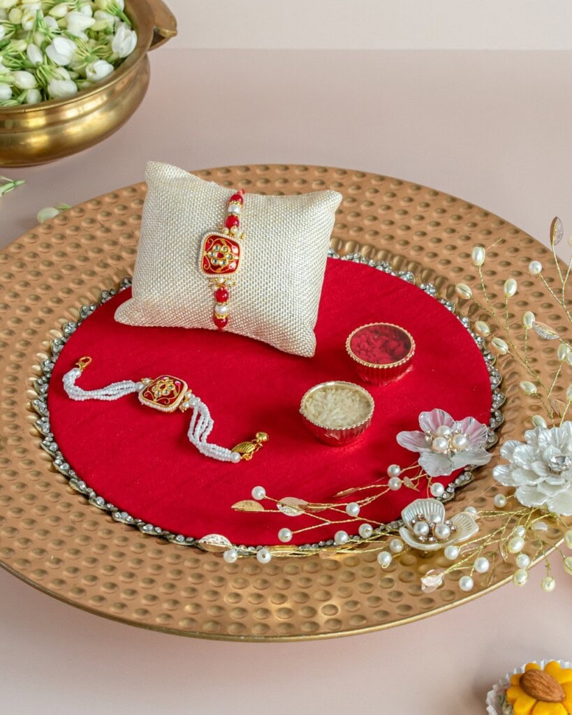 Floral Art presents A decorative gold tray with a red base holds two rakhis, a small round box, a white flower, a beige cushion, and scattered white buds.