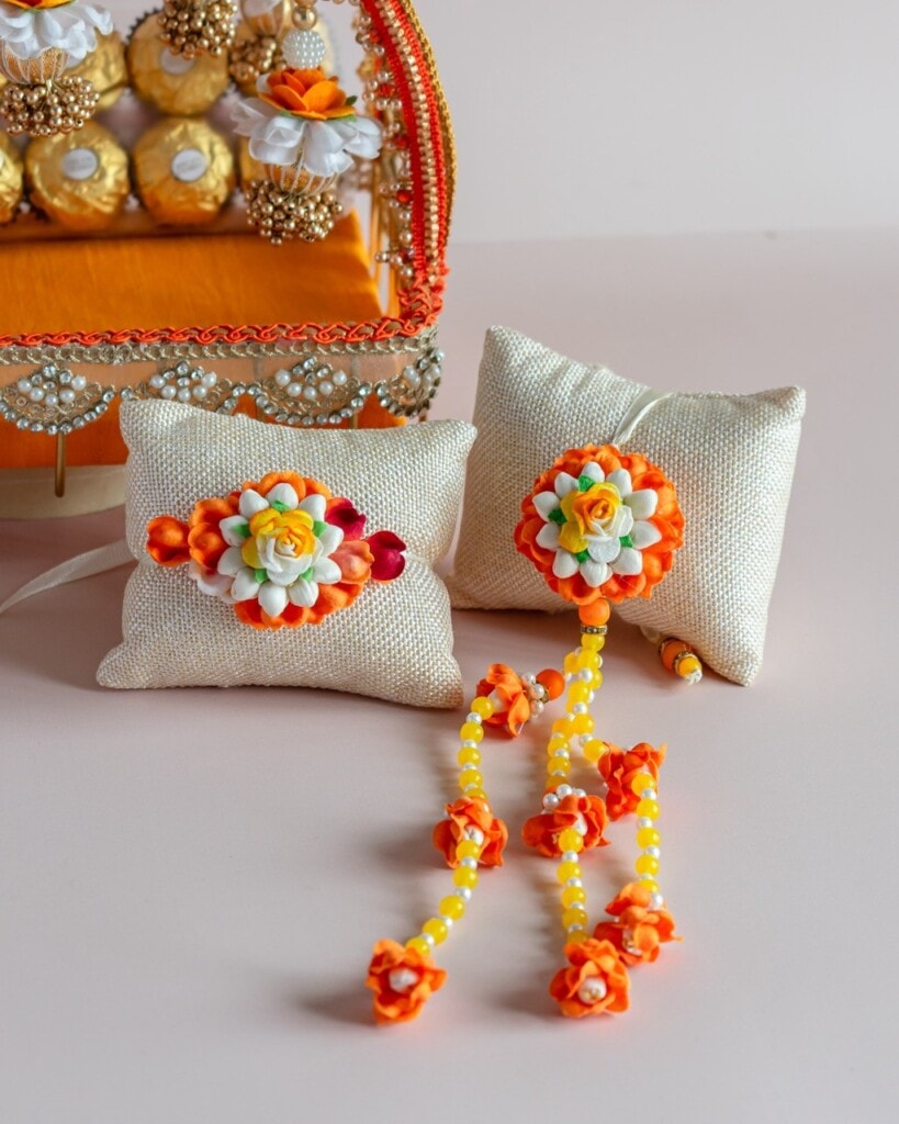 Floral Art presents Two decorative white cushions with orange, yellow, and white floral embellishments and beaded strings, placed on a light surface near a tray of wrapped chocolates.