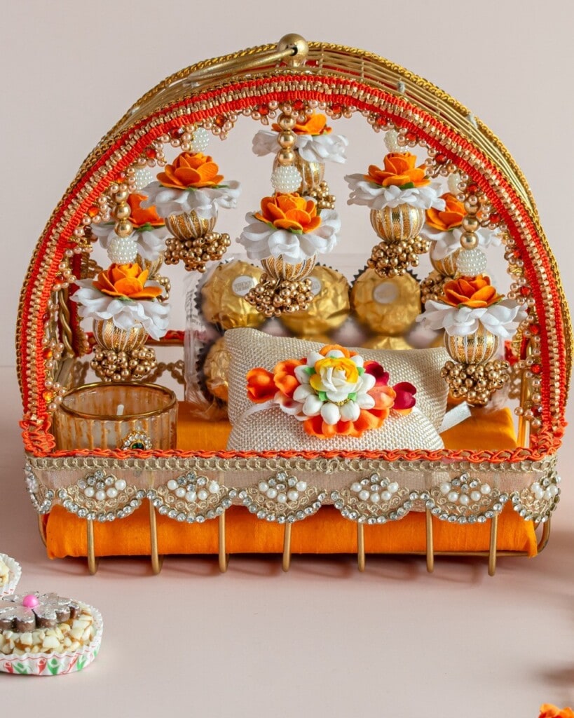 Floral Art presents An ornate decorative basket with orange and white flowers, pearls, and gold detailing, containing wrapped gifts and a glass, set on an orange cushion.