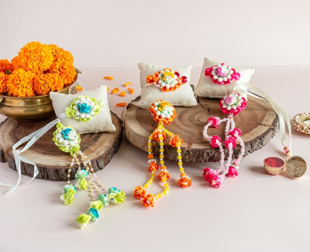 Floral Art presents Four decorative floral rakhis are displayed on wooden slabs alongside marigold flowers, cushions, and small containers on a light background.