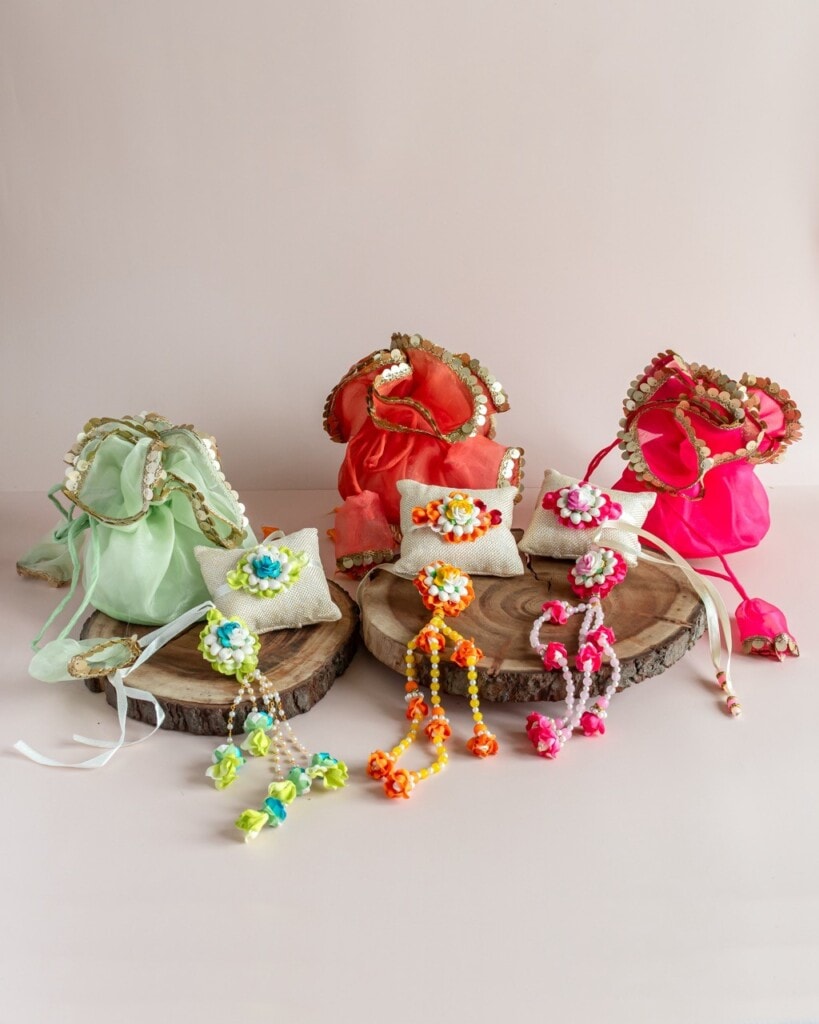 Floral Art presents Three decorative drawstring pouches in pastel green, orange, and pink are displayed with matching beaded accessories on wooden platforms against a pale pink background.