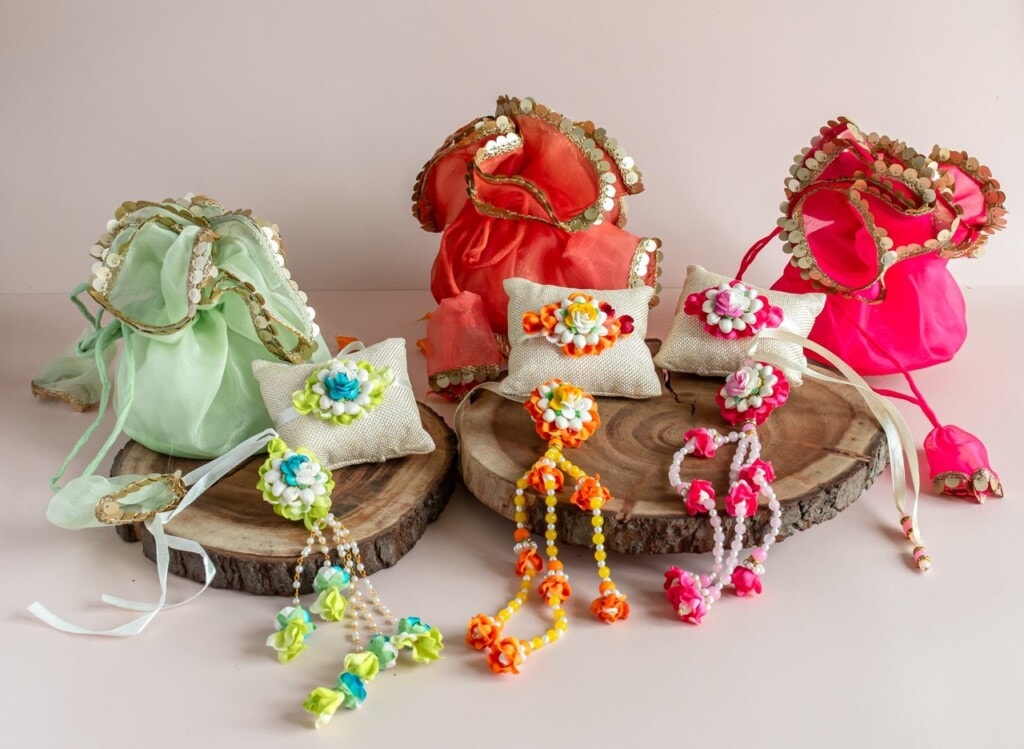 Floral Art presents Three fabric potli bags in green, coral, and pink with gold trim, and three ornamental rakhis with beads and flowers arranged on wooden slabs.