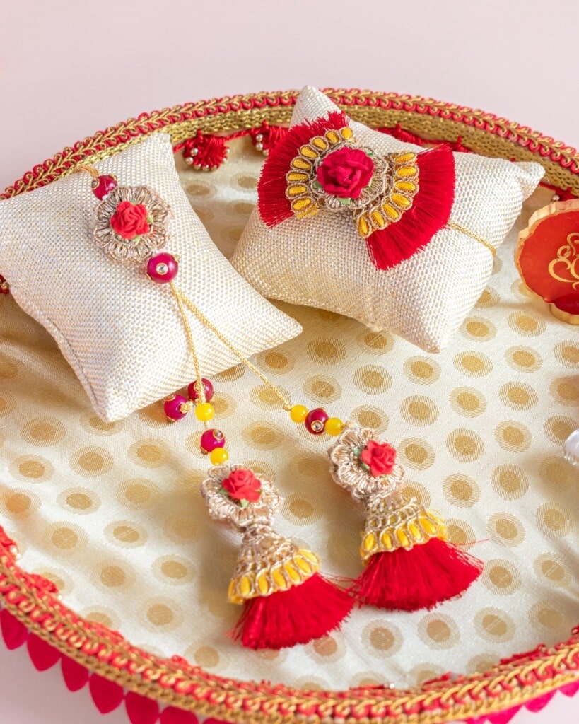 Floral Art presents A decorative tray with two white cushions displaying traditional jewelry with red and gold tassels, beads, and floral embellishments.