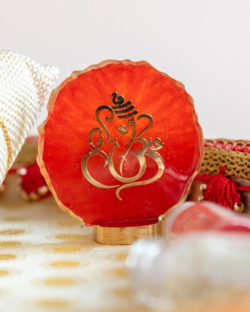 Floral Art presents A round decorative item with a red surface featuring a gold Ganesh symbol, placed on a patterned cloth with other festive objects nearby.