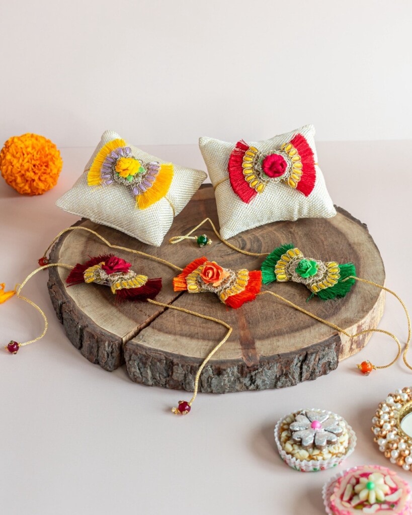 Floral Art presents Decorative rakhis with floral designs displayed on wooden slabs, accompanied by small cushions, a marigold flower, and festive sweets.