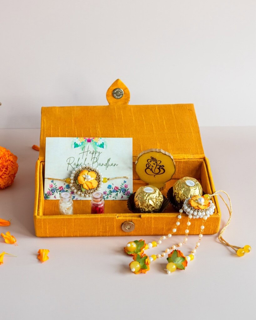 Floral Art presents A yellow gift box contains chocolates, a Rakhi bracelet, a greeting card, small bottles, and a decorative item with a Ganesh symbol, arranged on a light surface.