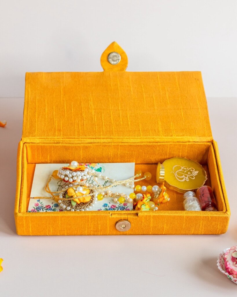 Floral Art presents A yellow fabric jewelry box holds floral jewelry pieces, a decorated envelope, and small containers, displayed on a light surface.