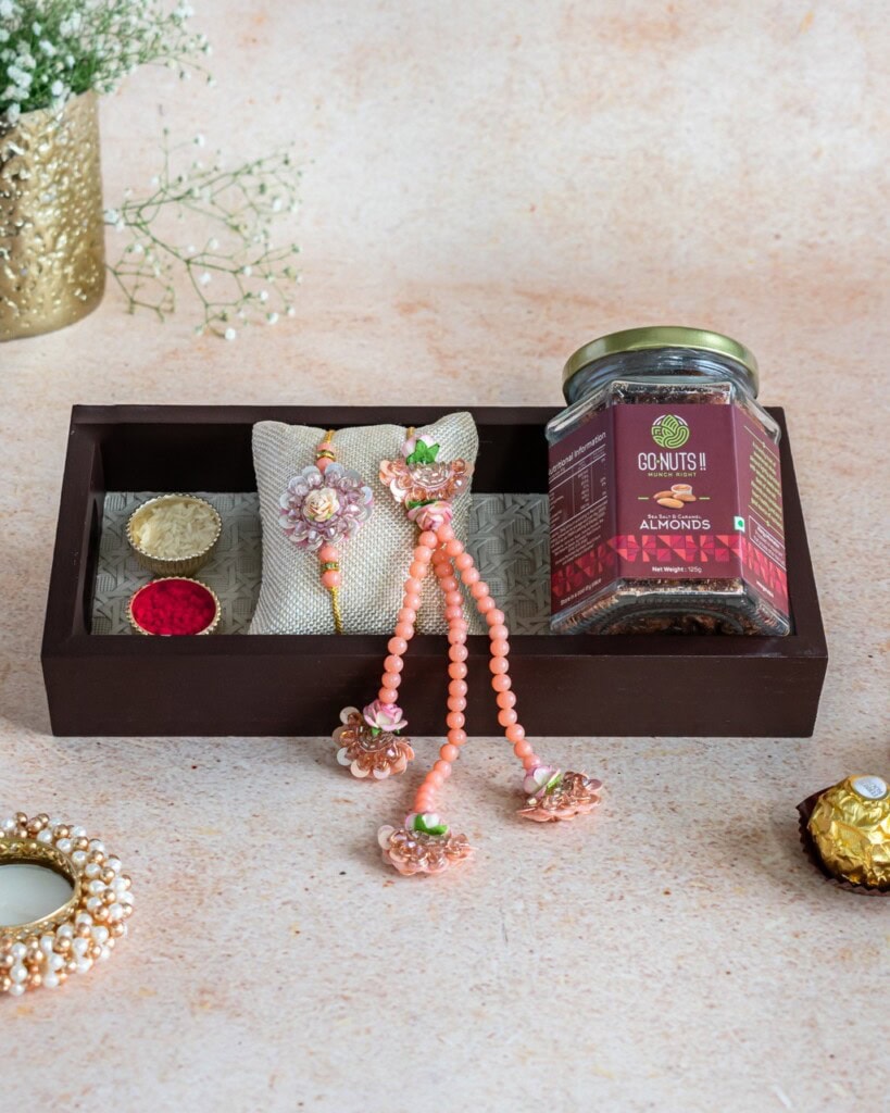 Floral Art presents A gift box contains a jar of almonds, decorative jewelry, a pair of round containers with red and white powder, and a Ferrero Rocher chocolate, arranged on a beige surface.