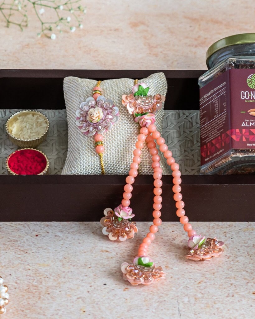 Floral Art presents A decorative peach floral rakhi and bracelet set displayed on a cushioned tray beside a jar of almonds and small bowls of rice and red powder.