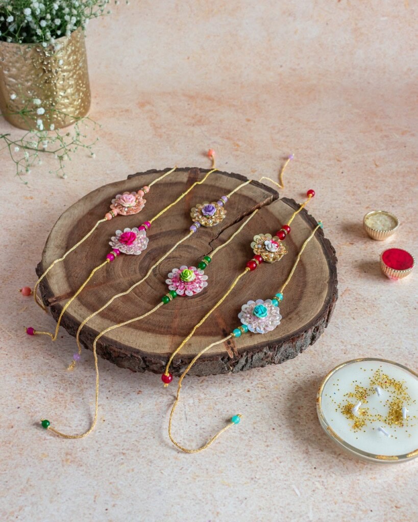 Floral Art presents Six decorative rakhis with colorful beads and floral designs are displayed on a wooden tray, with bowls of red powder and white liquid nearby.