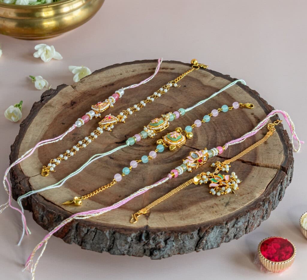 Floral Art presents Four decorative rakhis with beads and gold embellishments are displayed on a wooden slab, surrounded by flowers and small decorative containers.