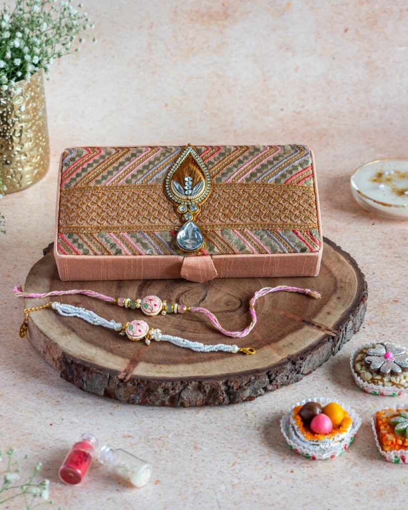 Floral Art presents An ornate clutch, two decorative rakhis, and traditional Indian sweets are arranged on a wooden slab with small decorative items and flowers nearby.