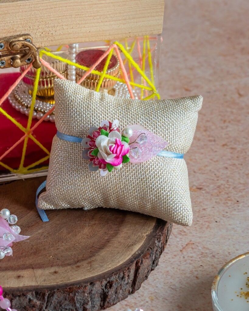 Floral Art presents A small burlap cushion decorated with a pink artificial flower and ribbon, displayed on a wooden surface with decorative boxes in the background.
