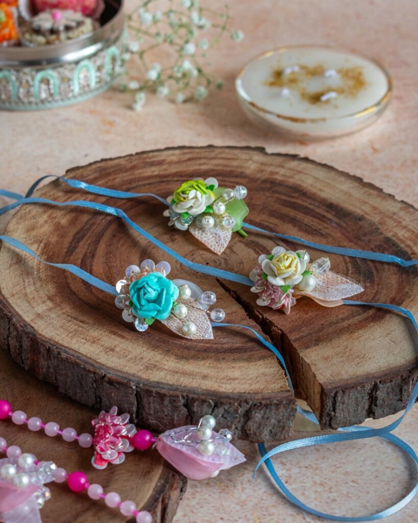 Floral Art presents Three decorative rakhi bracelets with floral and pearl designs are displayed on wooden slices, with additional decorative items in the background.