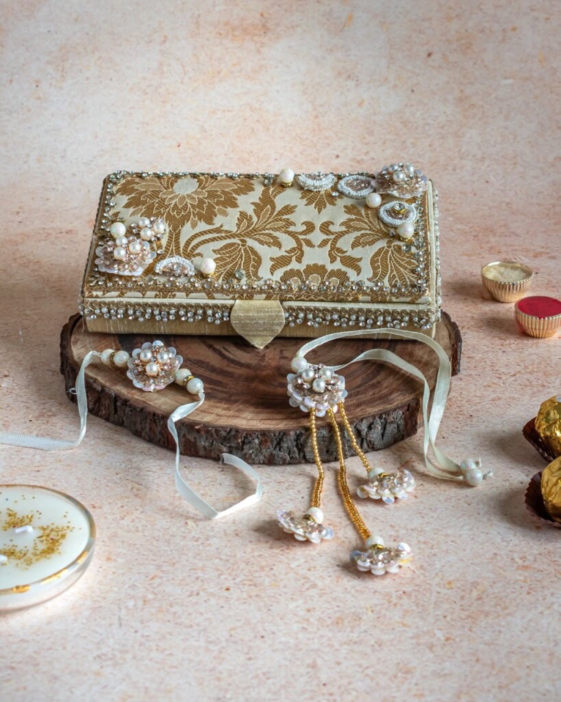 Floral Art presents A decorative clutch with gold floral embroidery and pearl embellishments is displayed on a wooden platform, surrounded by matching jewelry and sweets.