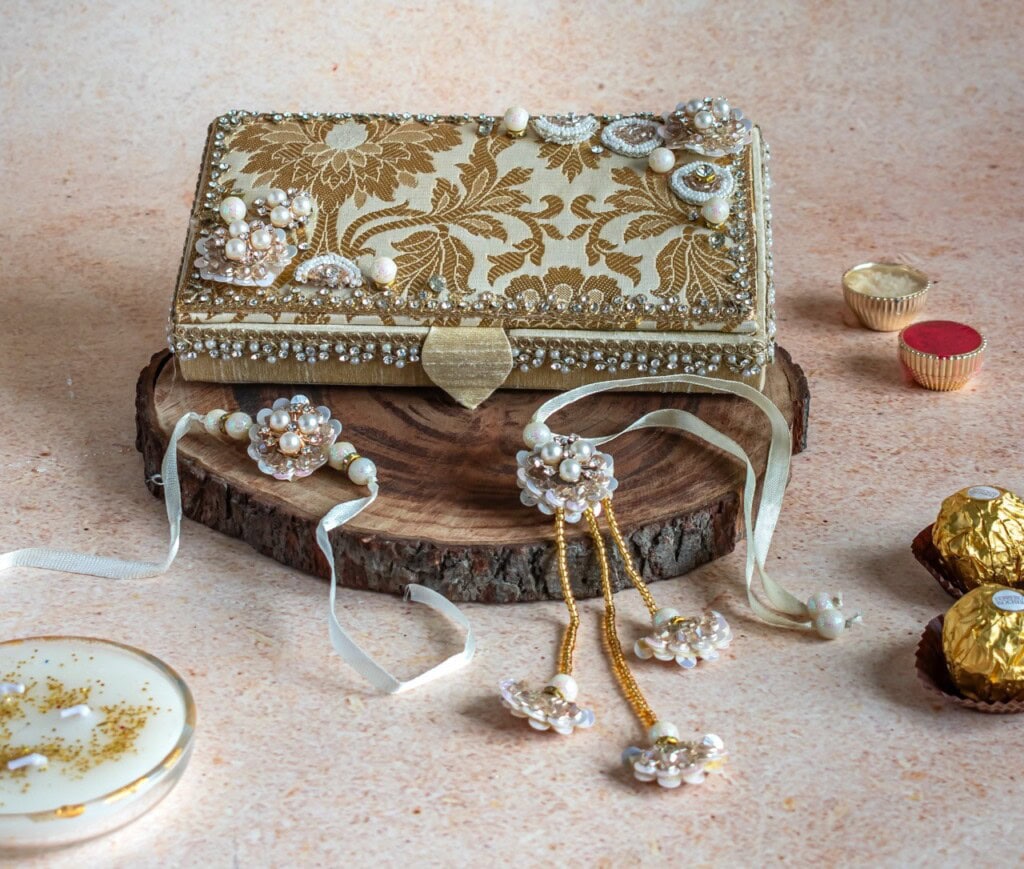 Floral Art presents A gold and beige embroidered clutch with pearl and bead embellishments sits on a wooden slab, surrounded by chocolates and decorative items.