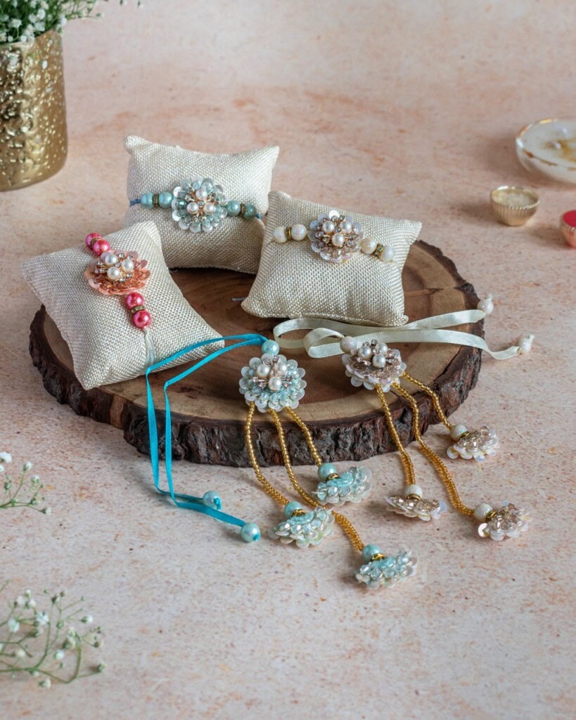 Floral Art presents Three decorative pearl and bead rakhis displayed on small fabric pillows, with additional matching accessories arranged on a wooden platter.