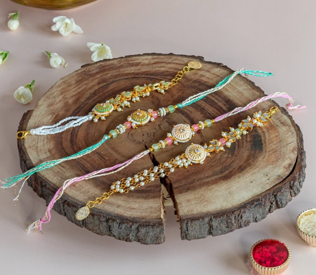 Floral Art presents Three decorative rakhi bracelets with beads and gold toned accents are displayed on a wooden slab, surrounded by flowers and festive items.