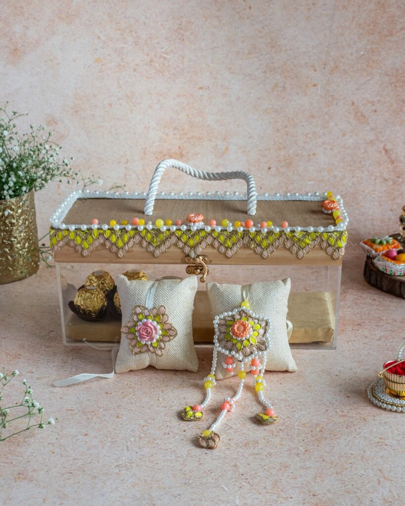 Floral Art presents A decorative box with floral beadwork, two embroidered cushions, and a beaded ornament are displayed on a textured surface with small flower arrangements nearby.