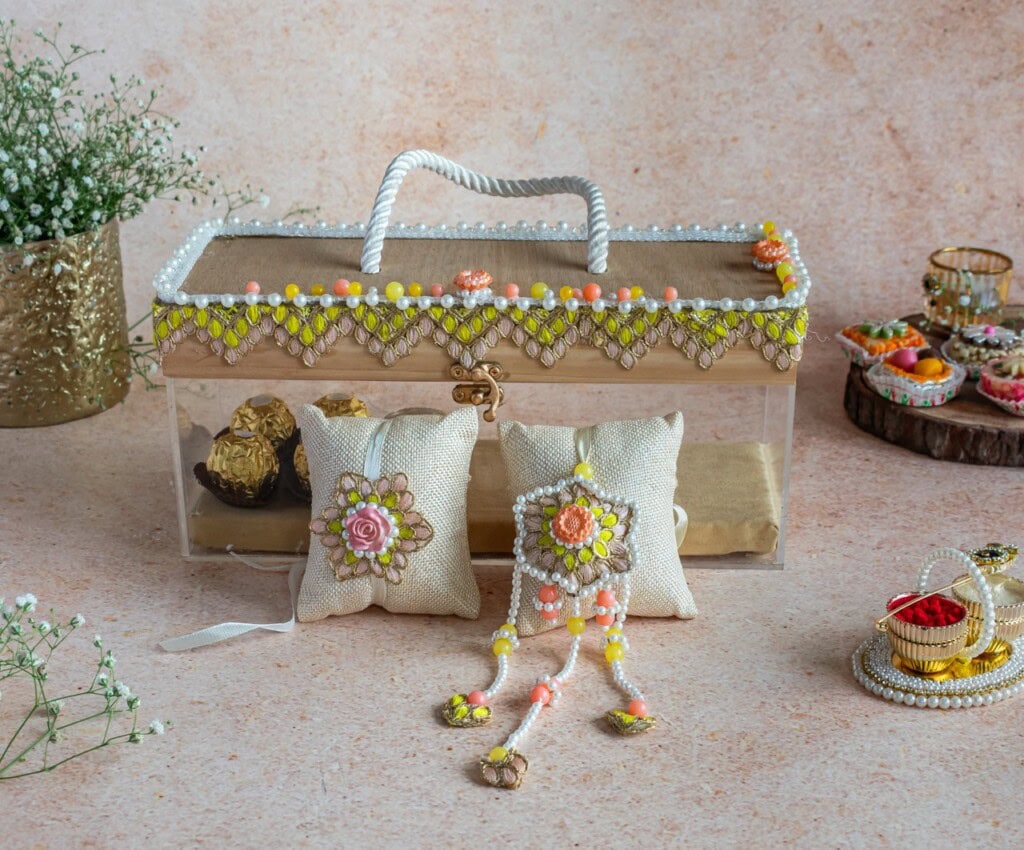 Floral Art presents A decorative transparent gift box with a beaded handle, two embroidered cushions, a beaded ornament, and trays with jewelry and flowers, arranged on a beige surface.