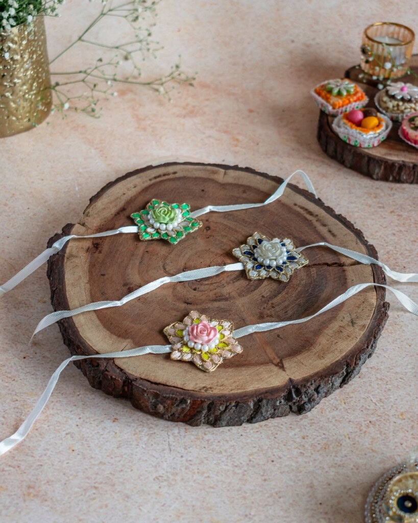 Floral Art presents Three decorative rakhis with floral designs and white ribbons are displayed on a round wooden tray, with festive sweets and decorations in the background.