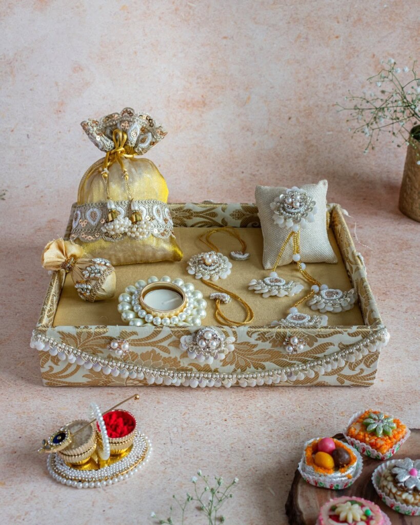 Floral Art presents A decorative tray with pearl and gold accents holds jewelry, a small pouch, a ring box, and festive sweets, set on a textured surface with scattered flowers.