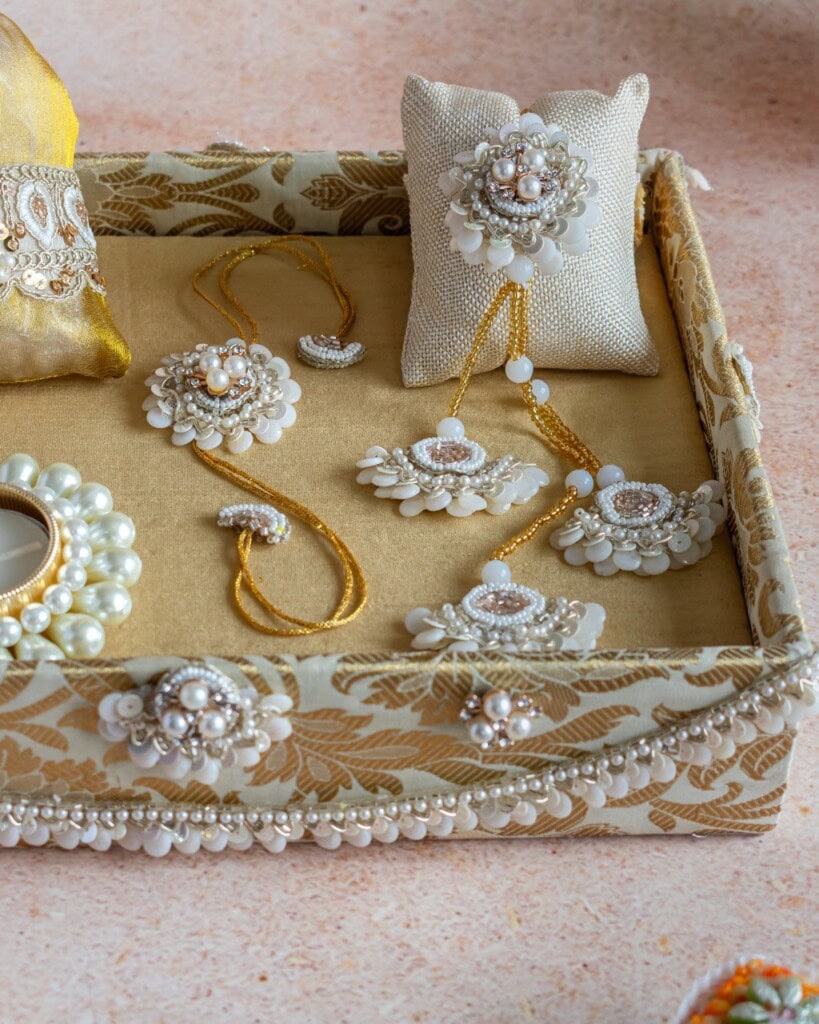 Floral Art presents A decorative box displaying pearl and bead jewelry pieces, including earrings, a bracelet, a ring, and a necklace, arranged on a beige fabric background.