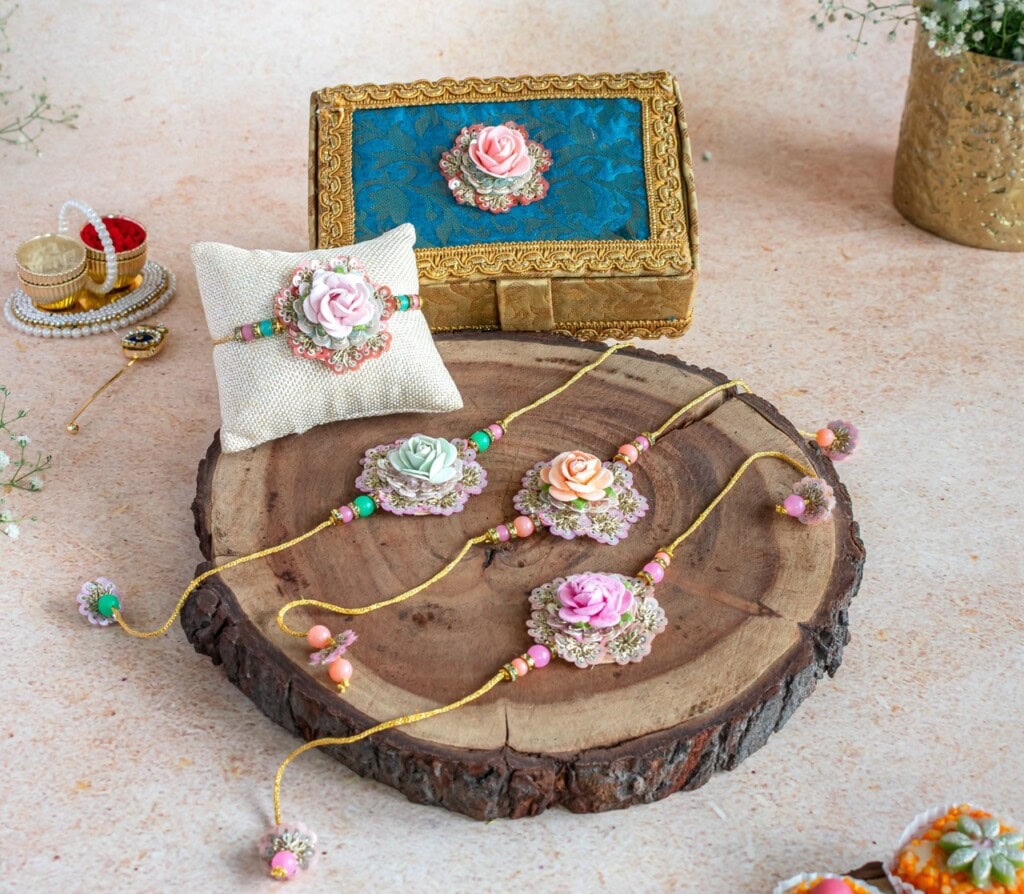 Floral Art presents Floral rakhis with decorative beads and artificial flowers displayed on a wooden slab, next to a cushion, ornate box, and small decorative items.