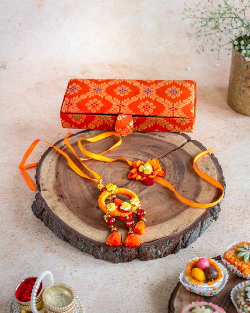 Floral Art presents An orange and yellow floral rakhi with beads and tassels is displayed on a wooden slab, with an ornate orange gift box and festive decorations around it.