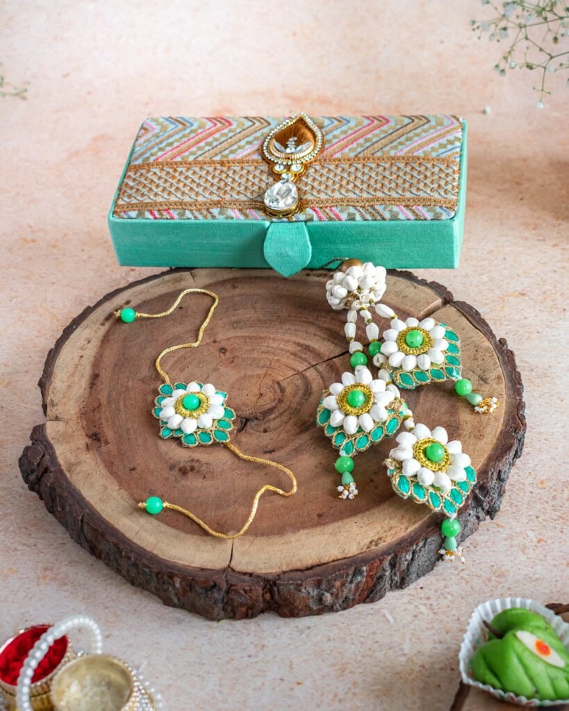 Floral Art presents A set of floral jewelry with green and white accents arranged on a wooden slab, next to a decorative mint green box and small bowls with red and white items.