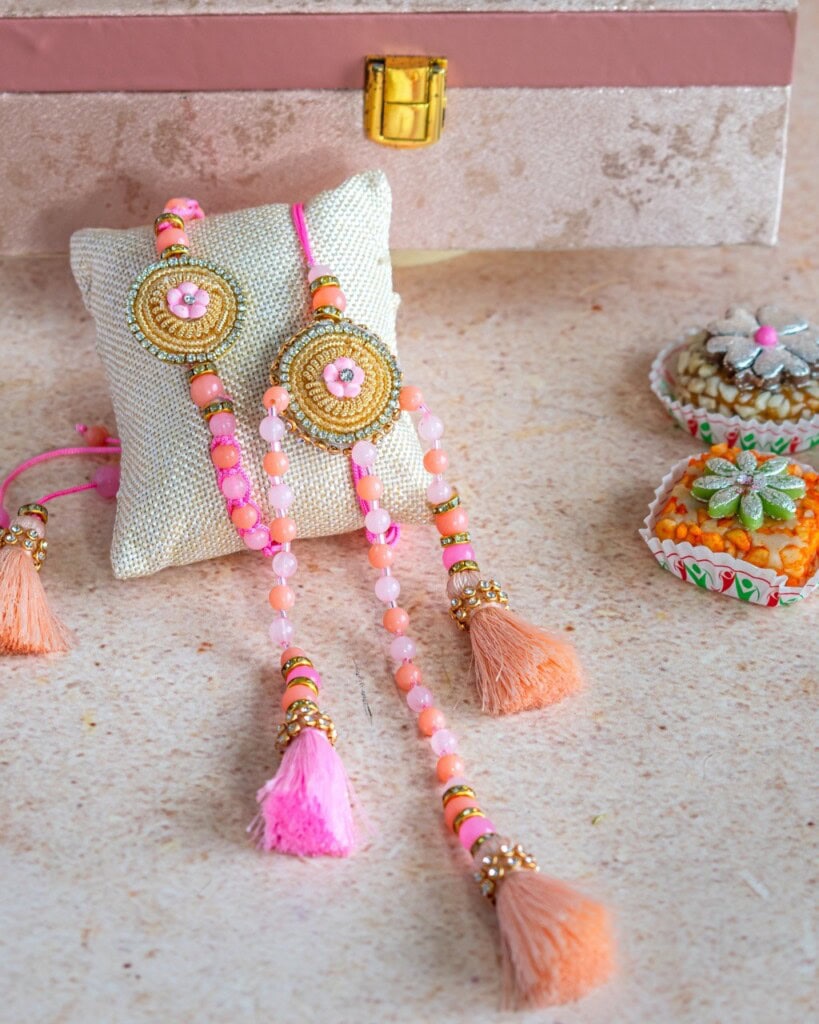 Floral Art presents Two decorative rakhi bracelets with pink and peach beads and tassels are displayed on a cushion, next to a decorative box and small bowls with festive items.
