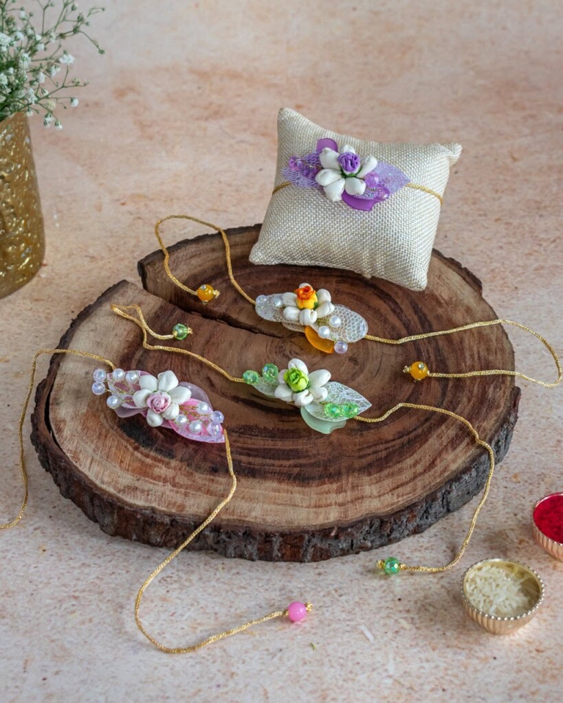 Floral Art presents Three decorative floral rakhis and a cushion are displayed on a wooden slab, with small bowls of rice and kumkum nearby, and a vase with white flowers in the background.