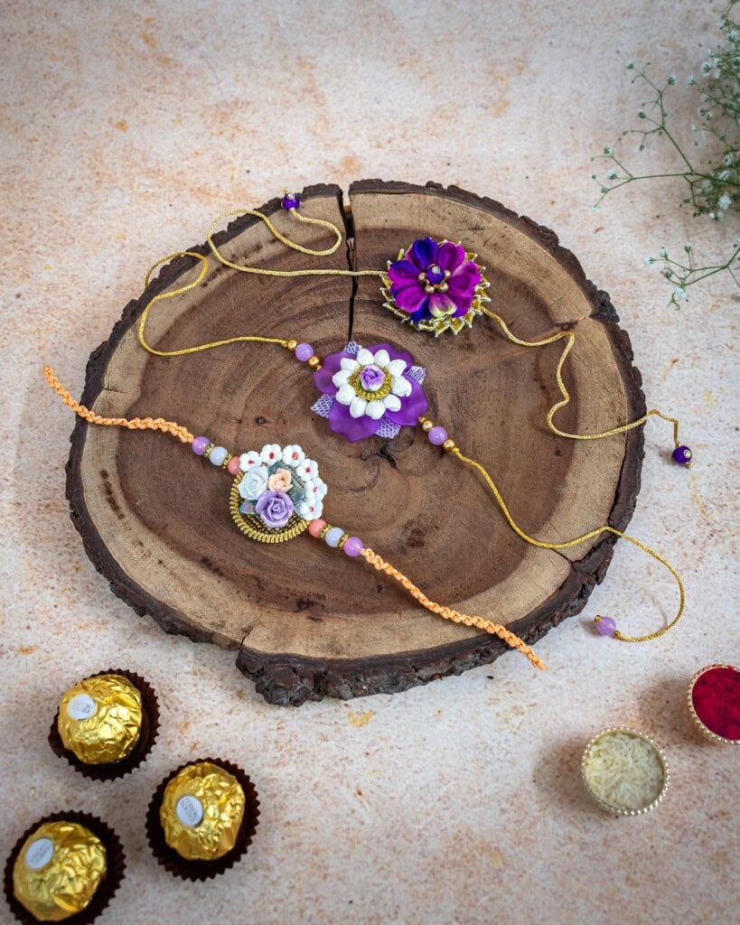 Floral Art presents Three decorative rakhis displayed on a wooden slab, with assorted sweets and flowers placed nearby on a light textured surface.
