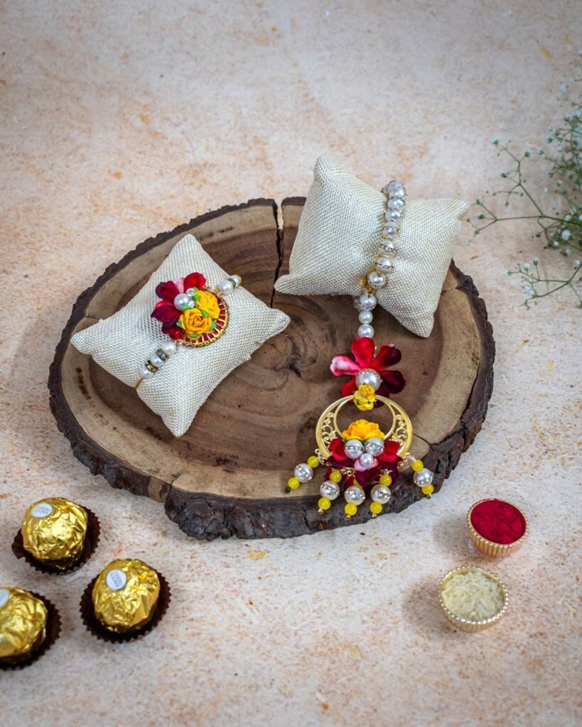 Floral Art presents A decorative rakhi and bracelet with red, yellow, and white flowers on cream cushions, placed on a wooden slab with chocolates and a small red powder container nearby.