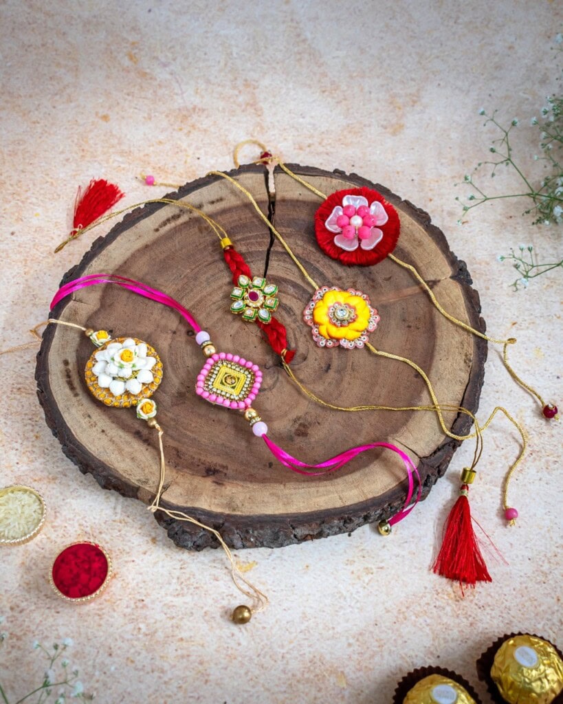 Floral Art presents Handcrafted decorative rakhis with colorful beads and tassels are arranged on a round wooden slab, with small flowers and red containers nearby.