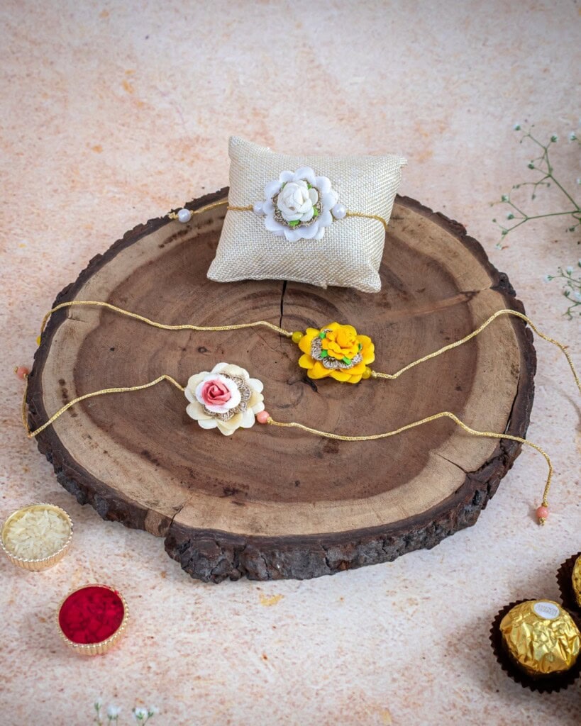 Floral Art presents Three decorative rakhis with floral designs are displayed on a wooden slab, along with a small pillow, sweets, and a bowl of red powder on a beige surface.
