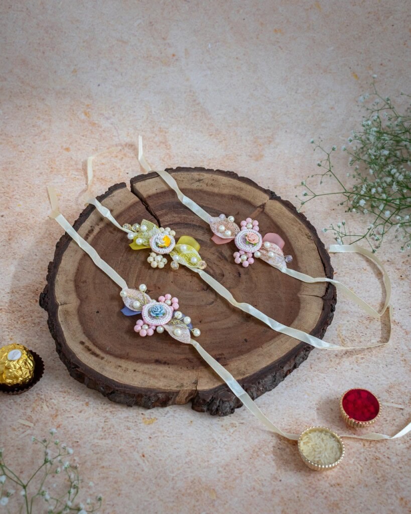 Floral Art presents Three decorative rakhis with pastel beads and ribbons are displayed on a wooden slab, surrounded by flowers and traditional Indian sweets on a textured surface.