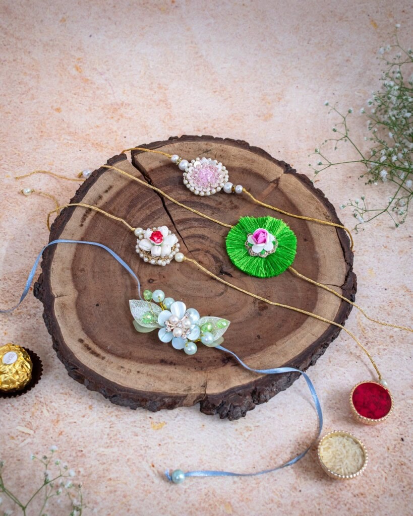 Floral Art presents Four decorative rakhis with different designs are displayed on a round wooden platter, with small flowers and gift containers nearby on a textured surface.