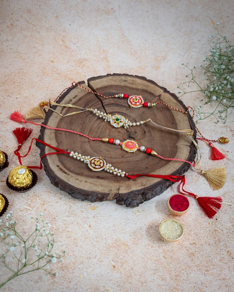 Floral Art presents Five decorative rakhis are arranged on a wooden slab, surrounded by flowers and traditional sweets on a light beige surface.