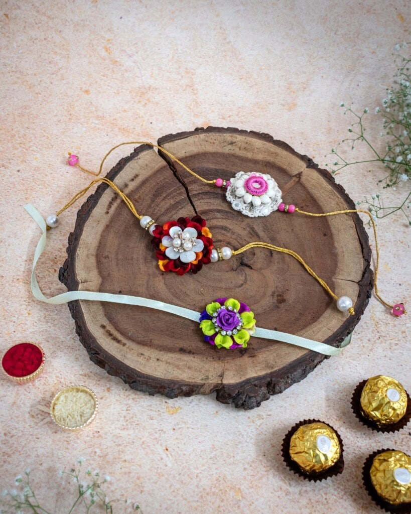 Floral Art presents Three decorative rakhis are displayed on a wooden slab, surrounded by assorted chocolates and small flowers on a light background.