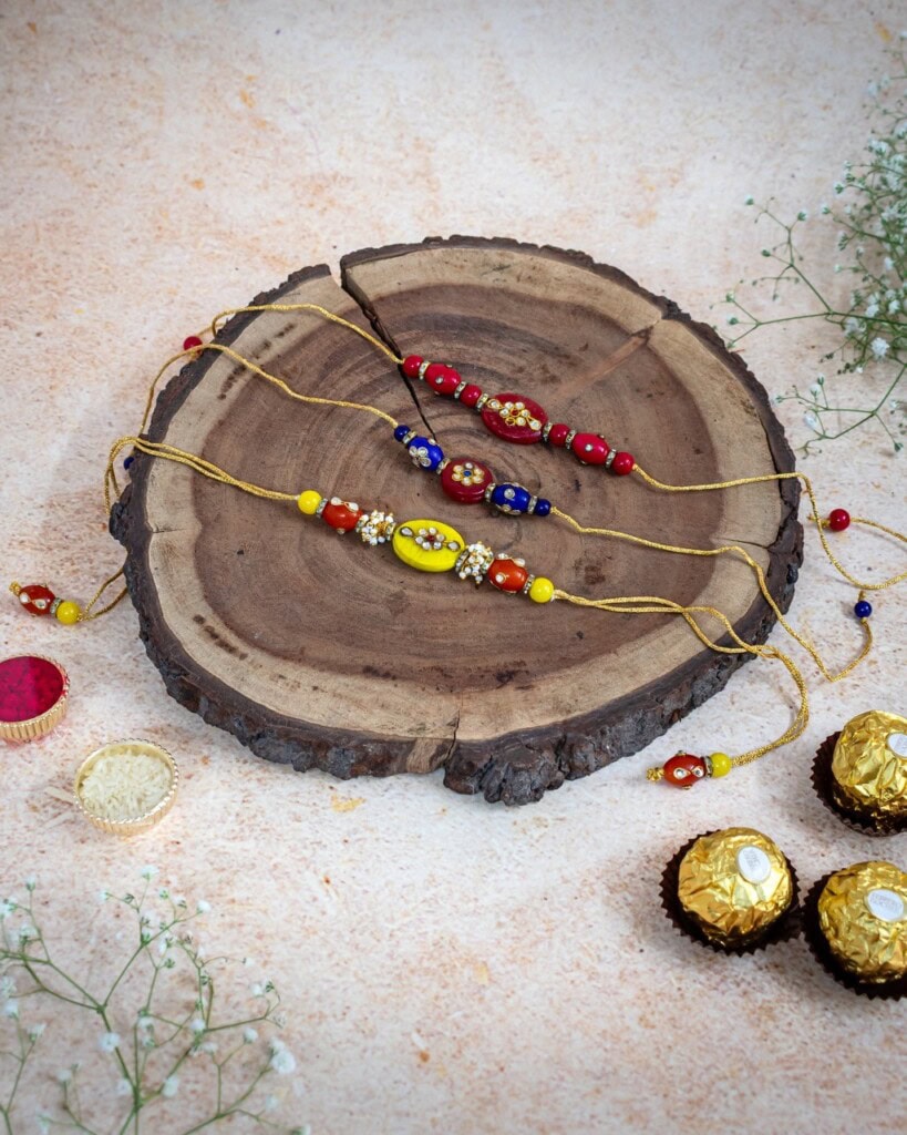 Floral Art presents Four decorative rakhis with red, yellow, and blue beads are displayed on a wooden slab, surrounded by Ferrero Rocher chocolates and small white flowers.