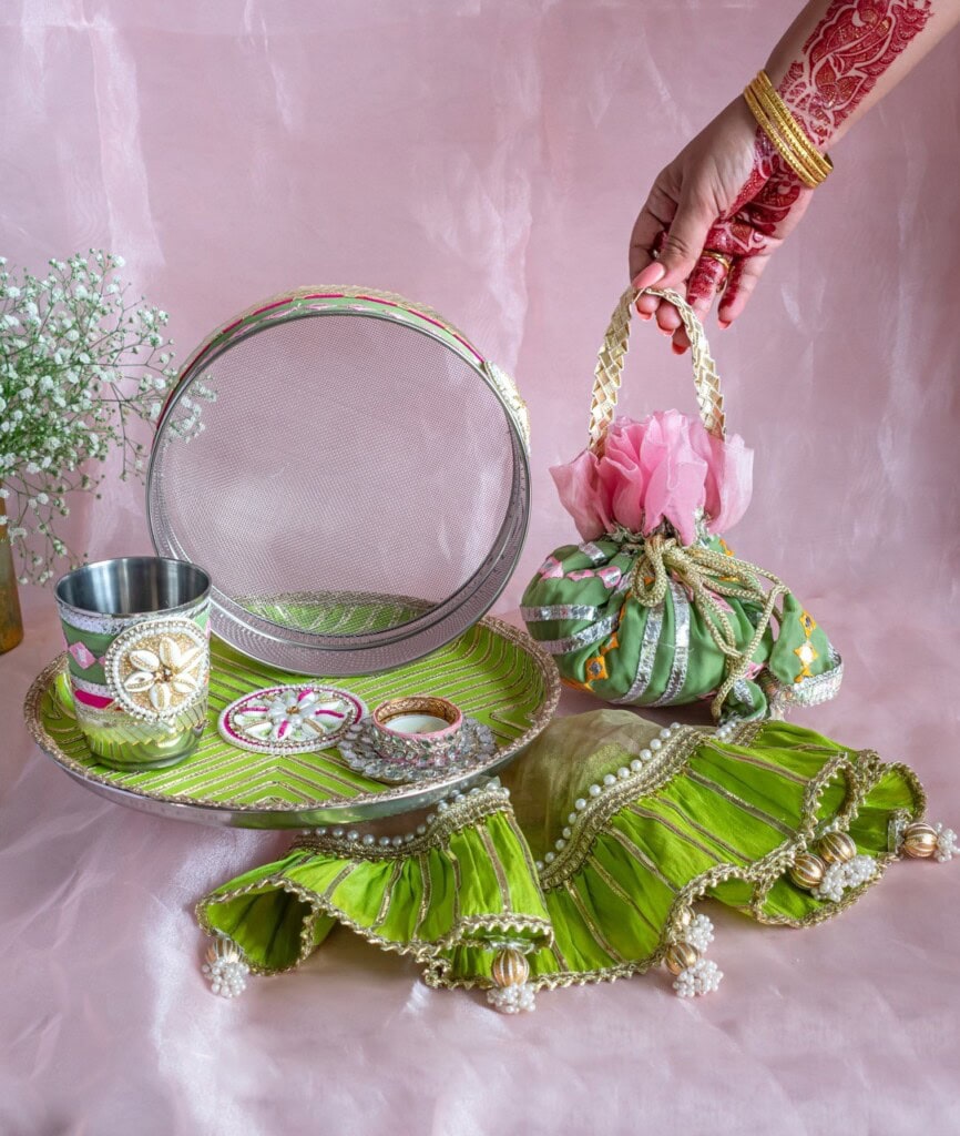 Floral Art presents Decorated traditional thali set with green fabric, metal glass, sieve, bangles, and pouch; a hand wearing red mehendi holds the pouch.