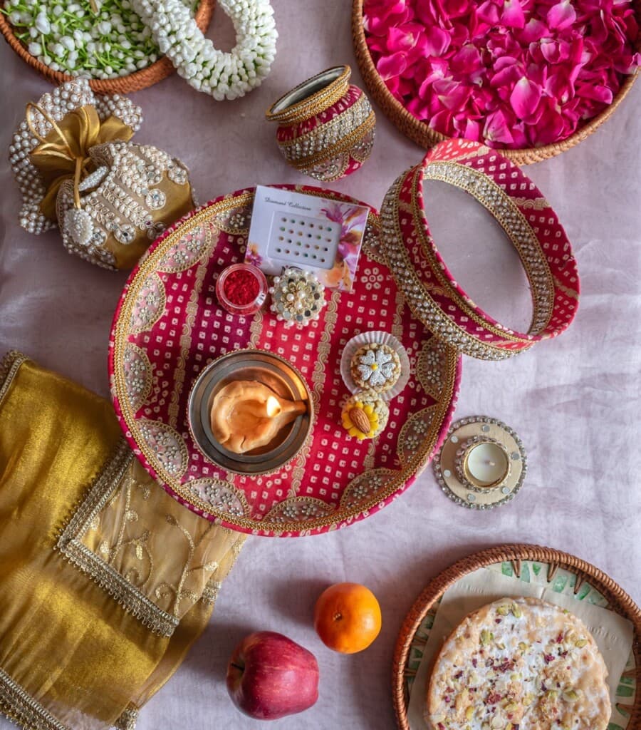 Floral Art presents A decorated thali with sindoor, diya, bangles, and sweets, alongside fruit, roti, rose petals, and flower garlands arranged for an Indian festival or ritual.
