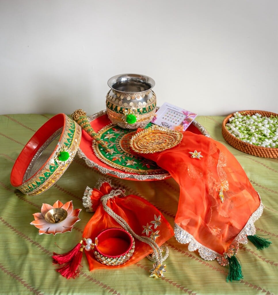 Floral Art presents Karva Chauth thali set with decorated sieve, pot, plate, sieve, red bangles, diya, prayer book, and orange dupatta, arranged on a green surface with a bowl of white flowers.