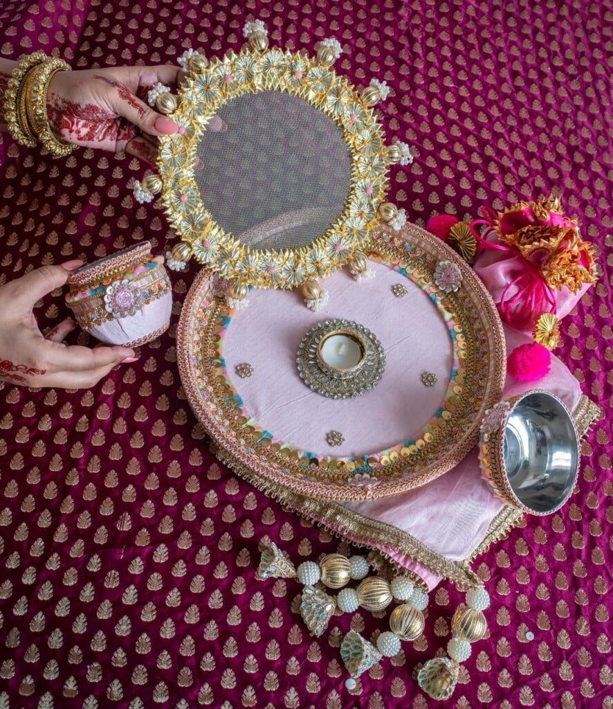 Floral Art presents A decorated thali set with a sieve, bowl, and cup is arranged on a maroon patterned cloth; a woman’s hands hold the sieve and cup.