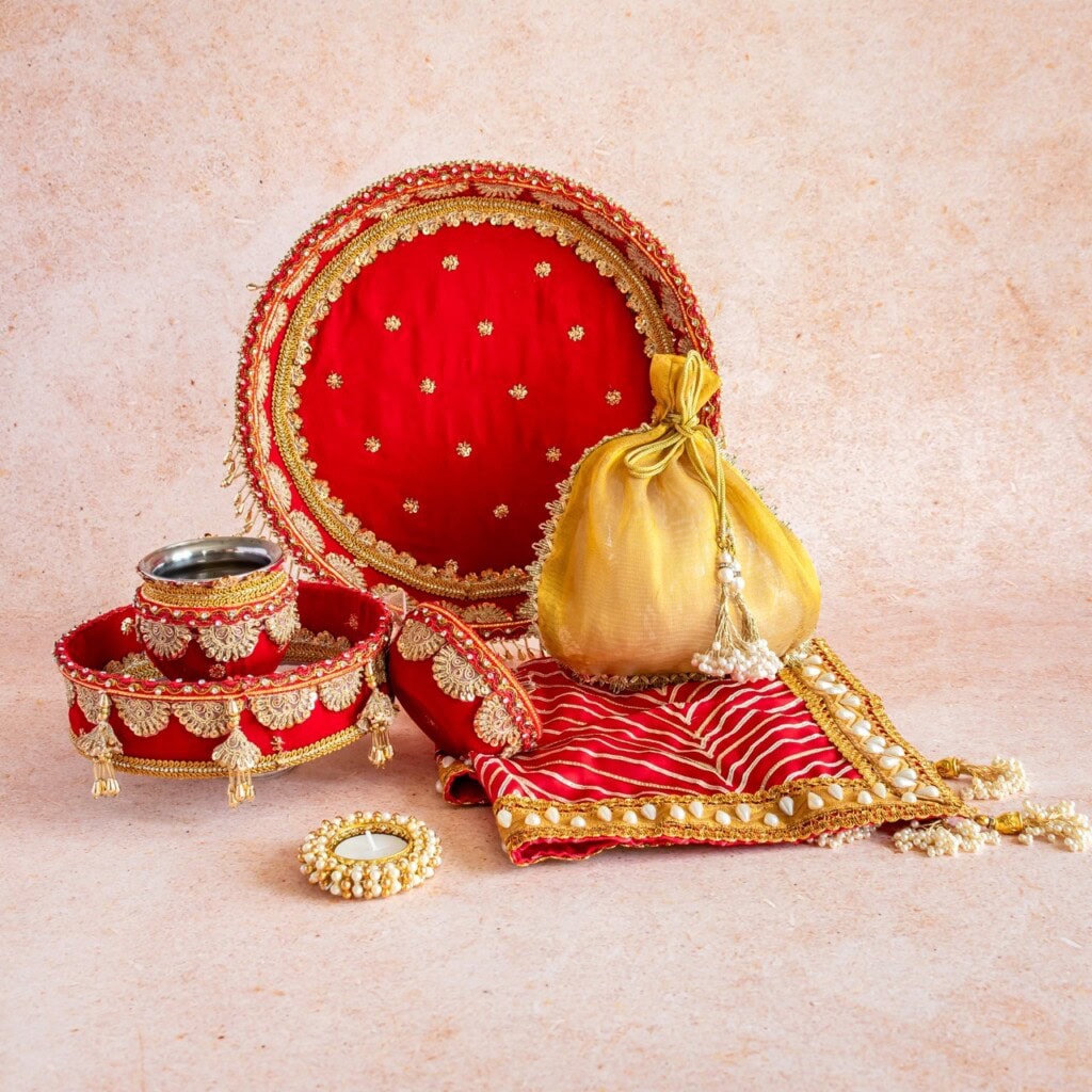 Floral Art presents A set of traditional Indian wedding items including a decorated red thali, gold potli bag, bangles, cloth, and tealight holder, all adorned with gold embroidery and embellishments.